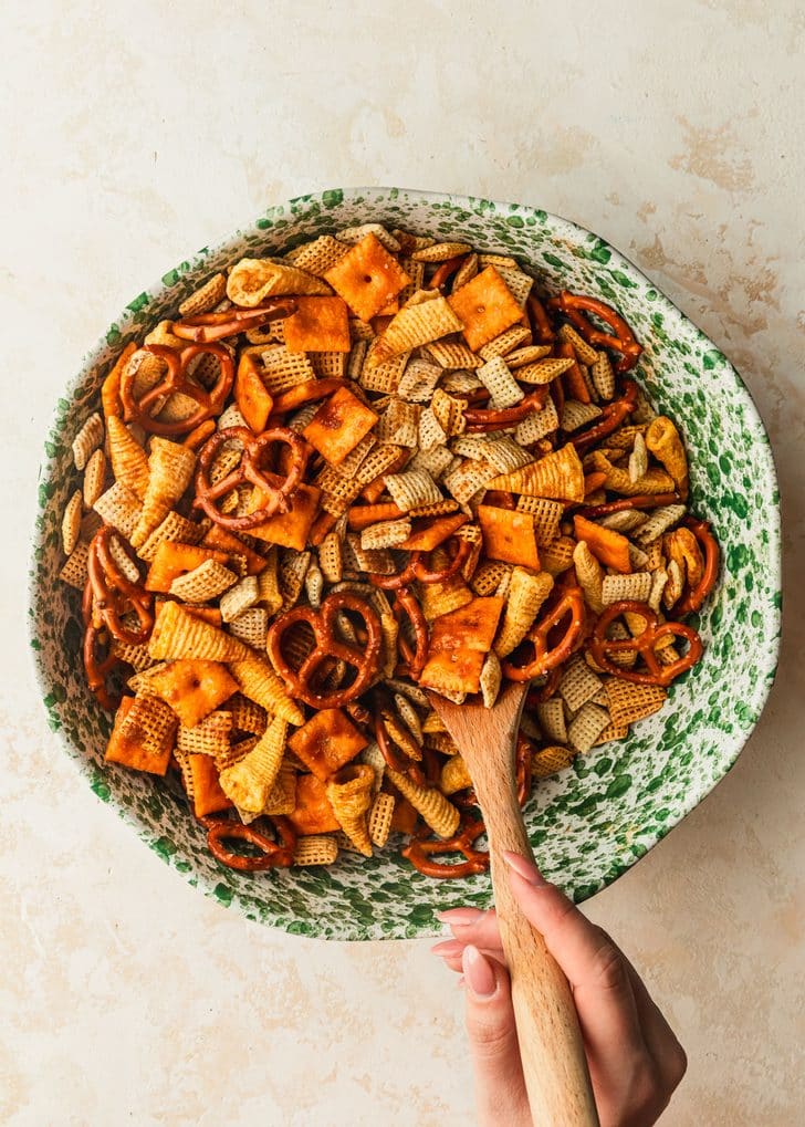 A hand using a wooden spoon to stir homemade cheddar cheese Chex mix in a green and white speckled bowl.