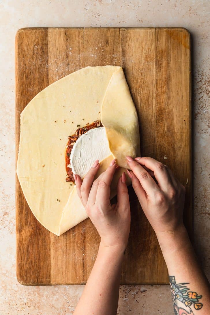 Hands folding brie in puff pastry for pumpkin-shaped baked brie.