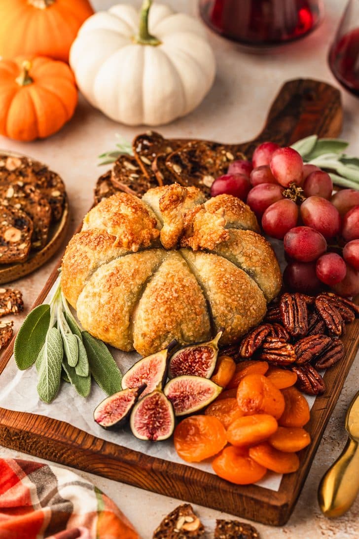 A wooden board topped with a pumpkin-shaped baked brie, pecans, crackers, grapes, and figs next to pumpkins, glasses of red wine, and a plaid linen on a brown marble counter.
