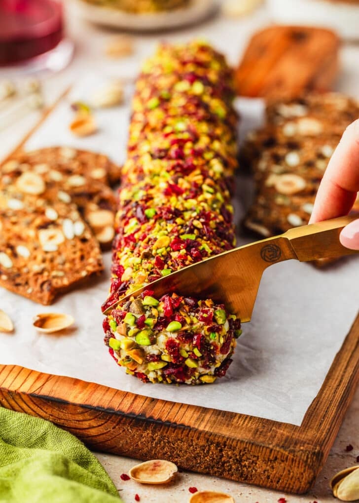 A hand using a cheese knife to slice a cranberry feta cheese log on a wooden board next to a glass of red wine, a brown bowl of pistachios, and green linen on a tan counter.