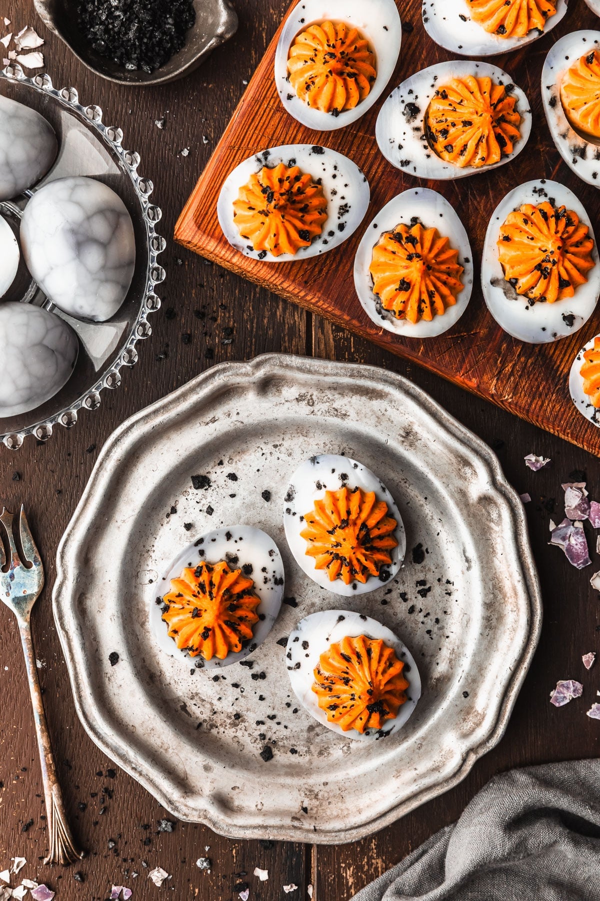 A silver plate and wooden platter of Halloween deviled eggs with a spider web design next to a grey linen, silver bowl of black salt, and clear plate of eggs on a wooden table.