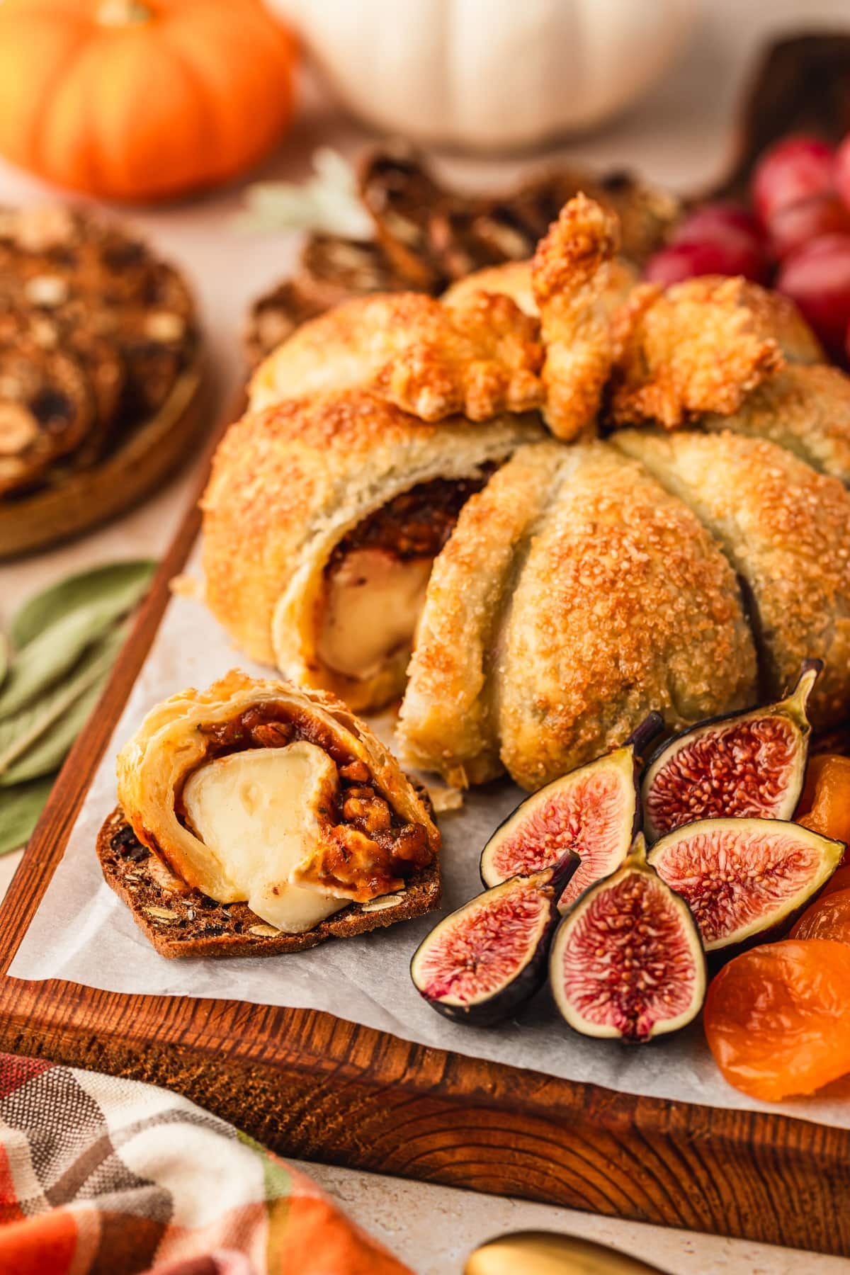 A sliced pumpkin-shaped baked brie with figs and grapes on a wooden cheese board next to pumpkins, a brown plate of crackers, and a plaid linen on a brown counter.