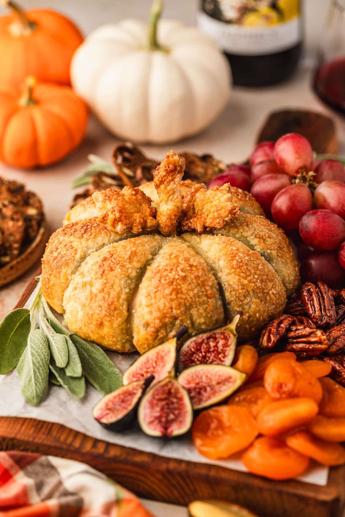 A pumpkin-shaped baked brie on a wood board with grapes, figs, and candied pecans on a brown counter next to pumpkins and a plaid linen.