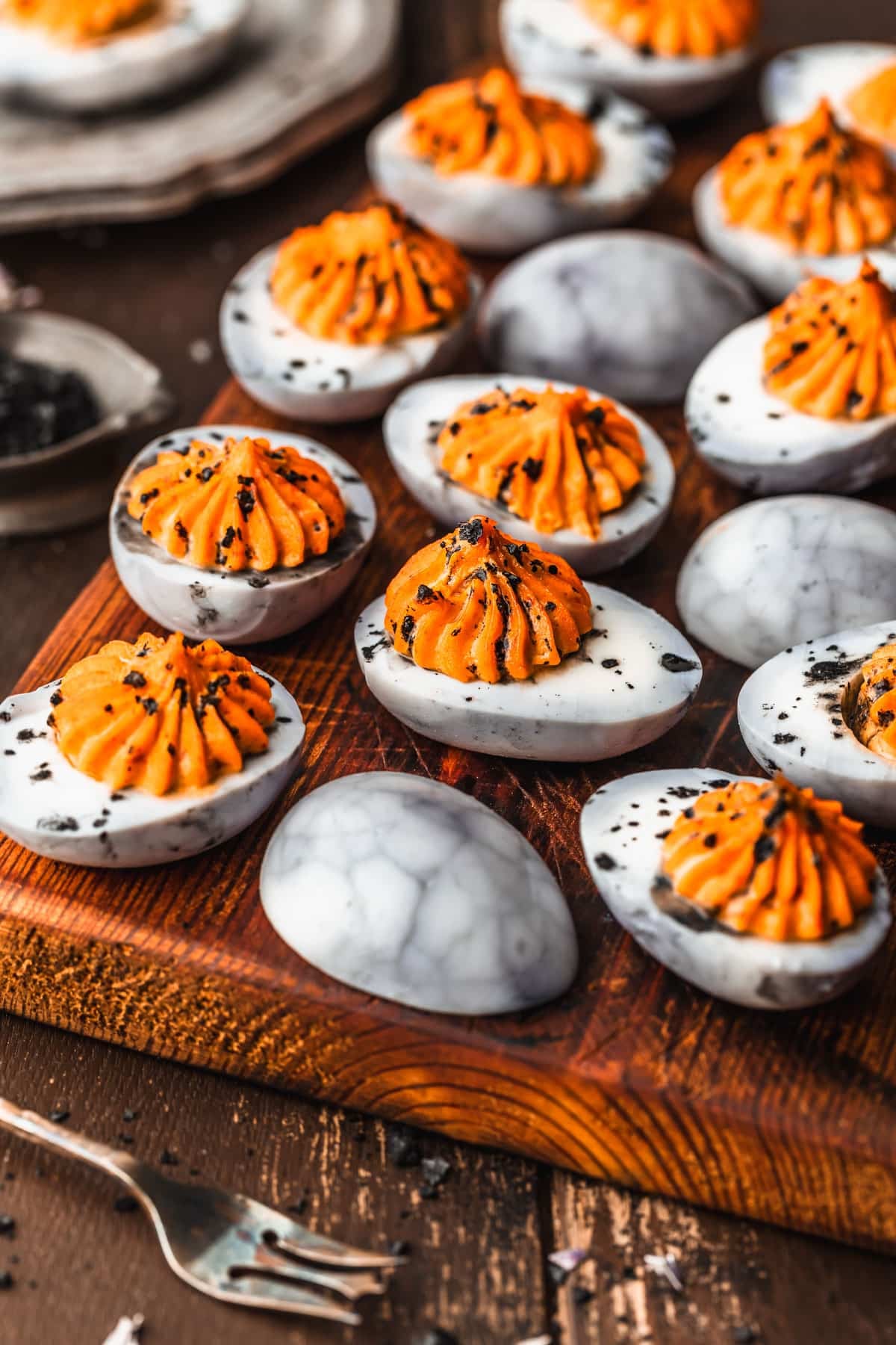 Rows of spider web Halloween deviled eggs on a wooden board next to a silver bowl of black salt, a silver plate, and a silver fork.